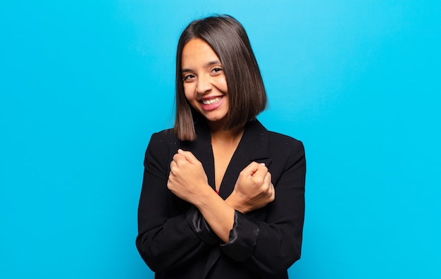 Mujer hispana sonriendo alegremente y celebrando, con los puños apretados y los brazos cruzados, sintiéndose feliz y positiva