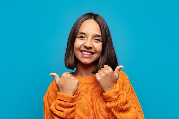 Mujer hispana sonriendo alegremente y luciendo feliz, sintiéndose despreocupada y positiva con ambos pulgares hacia arriba