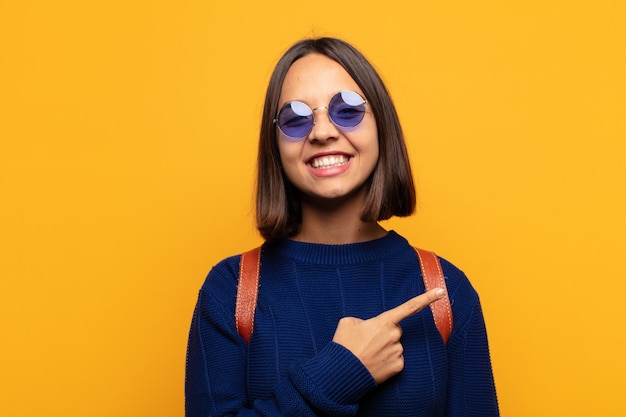 Mujer hispana sonriendo alegremente, sintiéndose feliz y apuntando hacia un lado y hacia arriba, mostrando el objeto en el espacio de la copia