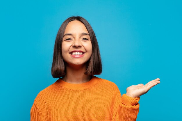 Mujer hispana sonriendo alegremente, sintiéndose feliz y mostrando un concepto en el espacio de la copia con la palma de la mano