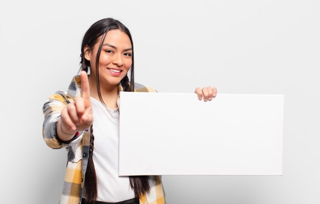 Mujer hispana sonriendo con orgullo y confianza haciendo la pose número uno triunfalmente, sintiéndose como una líder