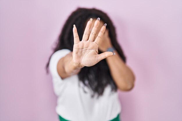 Mujer hispana de talla grande parada sobre fondo rosa cubriéndose los ojos con las manos y haciendo gestos de parada con expresión triste y de miedo concepto avergonzado y negativo