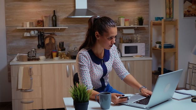 Mujer hispana con tarjeta de crédito en la cocina de casa para realizar pagos en línea. Empleado que realiza transacciones de pago desde casa en un portátil digital conectado a Internet trabajando horas extras para el trabajo