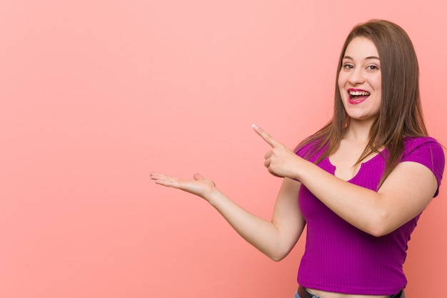 La mujer hispánica joven contra una pared rosada excitó sostener un espacio de la copia en la palma.