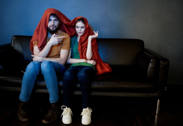 una mujer y un hombre debajo de una manta roja en el sofá viendo la televisión por la noche