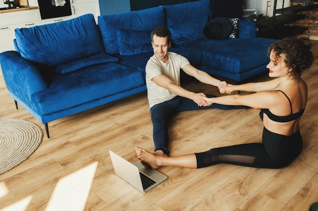 Mujer y hombre en forma en ropa deportiva haciendo yoga en el piso en casa usando una computadora portátil con tutorialsaludando a los estudiantesLos entrenadores masculinos tienen una clase virtual de yoga en la computadoraConcepto de estilo de vida activo