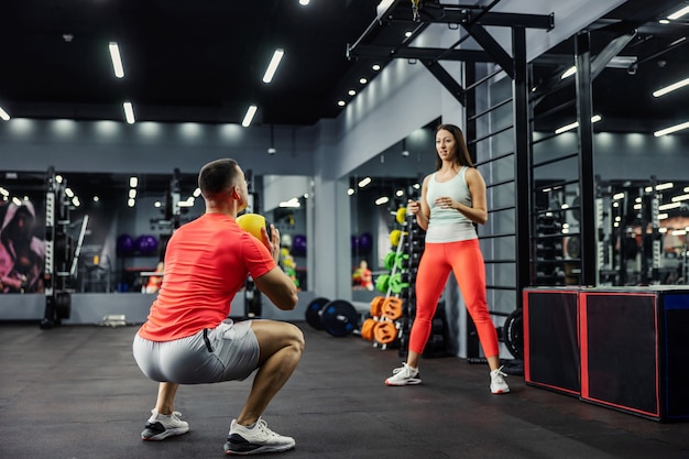 Una mujer y un hombre en ropa deportiva lanzan una pelota de fitness en el gimnasio. El hombre está en posición de cuclillas y se prepara para lanzar el balón a la chica que está de pie. Desafío deportivo, gol de pareja