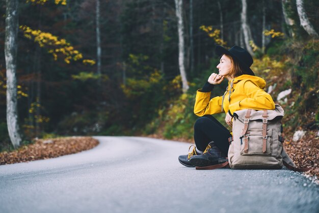 Mujer en impermeable amarillo caminando por la carretera en el bosque de otoño Chica joven excursionista en la naturaleza al aire libre