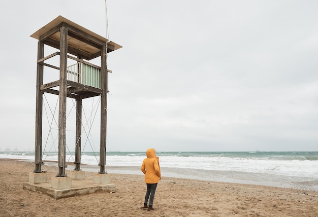 Mujer en impermeable amarillo mirando al mar. Camine por la playa en un día de mal tiempo.