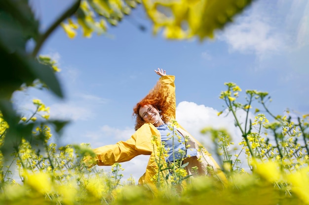 Una mujer con un impermeable amarillo saltando en el campo de flores amarillas y divirtiéndose en un día soleado