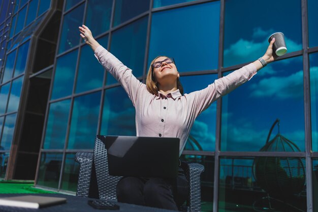 Foto mujer independiente alegre con una computadora portátil para la planificación del flujo de trabajo posando en un café callejero mujer exitosa con gafas con una computadora y una taza de café sonriendo al aire libre