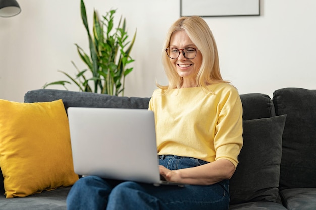 Una mujer independiente de alto nivel con un suéter amarillo sentada cómodamente en el sofá de la sala de estar escribiendo correos electrónicos en su portátil, blogueando y escribiendo mensajes.