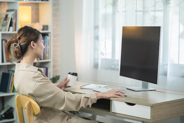 Mujer independiente asiática sonriendo sosteniendo una taza de café caliente y trabajando en una computadora portátil en una mesa de madera en casa Mujer emprendedora que trabaja para su negocio en casa Concepto de trabajo empresarial en casa