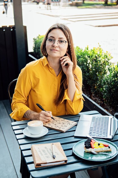 Mujer independiente escribiendo números en el bloc de notas mientras trabaja con una computadora portátil en un café callejero