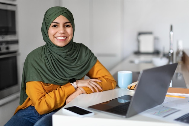 Mujer independiente del Medio Oriente en Hijab sentada en el escritorio con una computadora portátil