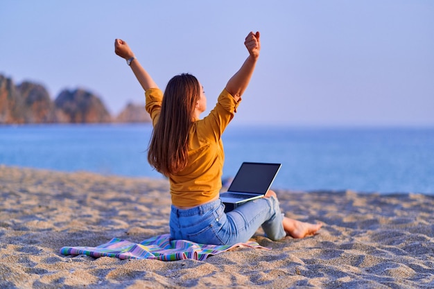 Mujer independiente milenaria satisfecha con los brazos abiertos usando la computadora y sentada en la playa de arena junto al mar Disfrute del concepto de trabajo remoto de la oficina de ensueño