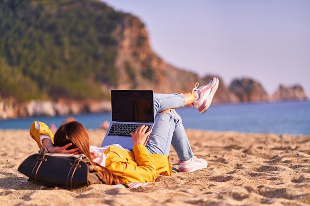 Mujer independiente milenaria usando una computadora portátil de maqueta con pantalla vacía en blanco y acostada en la playa de arena junto al mar Concepto de trabajo remoto de oficina de ensueño