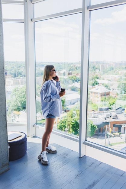 Mujer independiente moderna hablando por teléfono mientras está de pie en una ventana grande y bebiendo café El concepto de una mujer exitosa moderna Niña en una oficina abierta