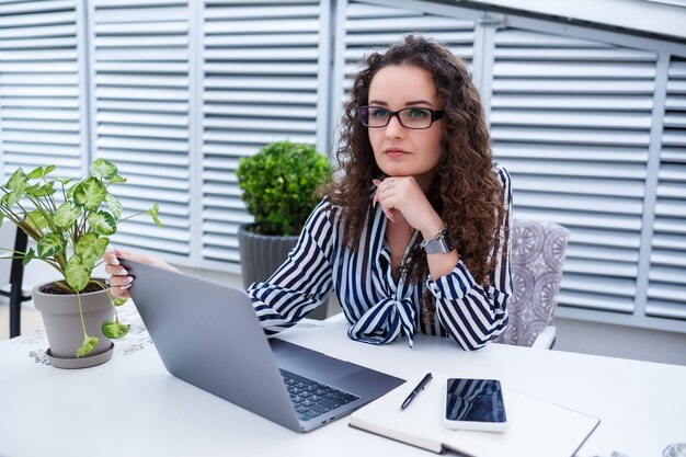 Mujer independiente pensativa en ropa casual de moda que trabaja con la computadora portátil y toma notas, sonriendo y mirando en la terraza al aire libre del café