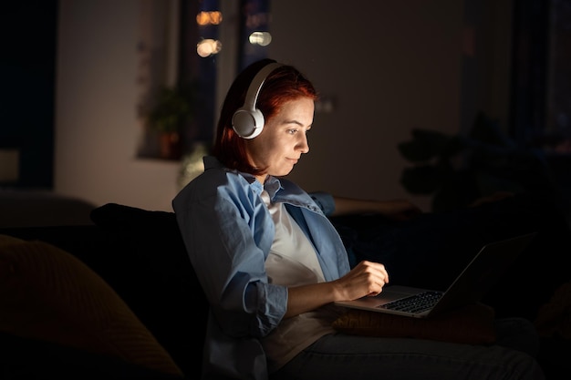 Mujer independiente positiva que trabaja con una computadora portátil en casa tarde en la noche escuchando música con auriculares