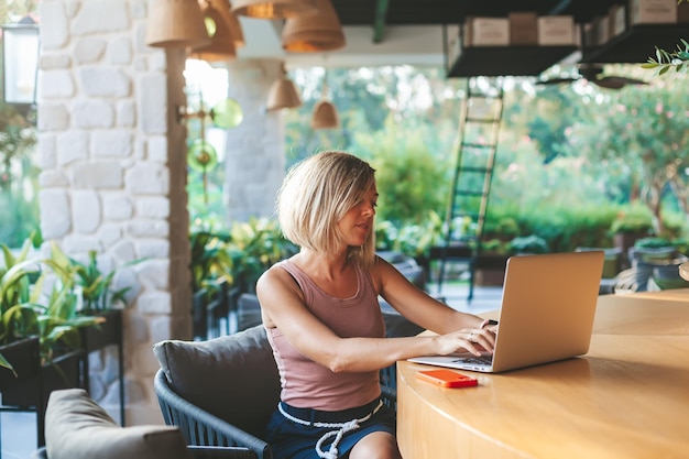 Mujer independiente que trabaja en el soleado café al aire libre usando una computadora portátil. Acogedor lugar de trabajo de oficina, trabajo remoto y concepto de trabajo en casa.