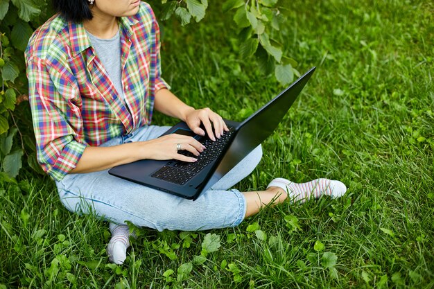 Mujer independiente en ropa casual con su computadora portátil sentada en el césped en el parque, trabajo independiente con notebook, oficina inteligente al aire libre, niña estudiando, educación en línea. Copia espacio
