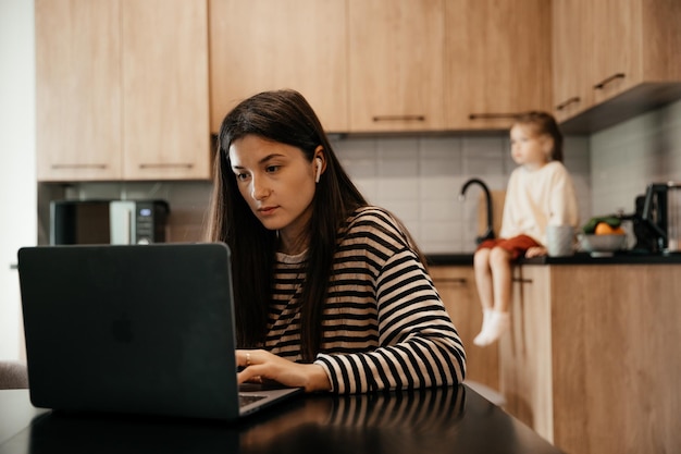 Mujer independiente sentada junto a la mesa en la oficina de la cocina del hogar trabajando en una computadora portátil Niño juguetón distrae del trabajo niño haciendo ruido y pidiendo atención de mamá ocupada