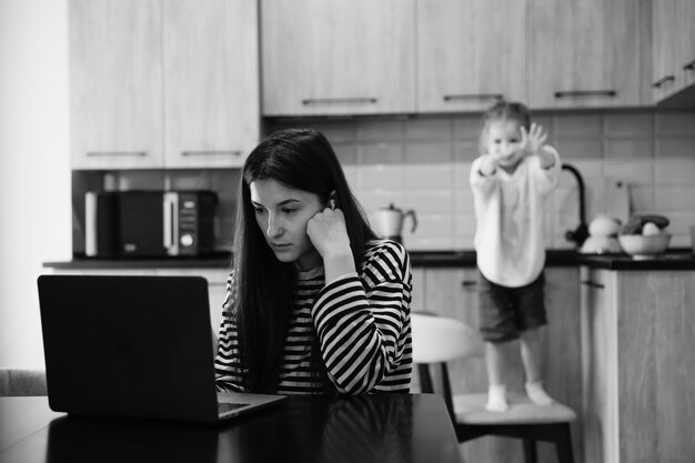 Mujer independiente sentada junto a la mesa en la oficina de la cocina del hogar trabajando en una computadora portátil Niño juguetón distrae del trabajo niño haciendo ruido y pidiendo atención de mamá ocupada