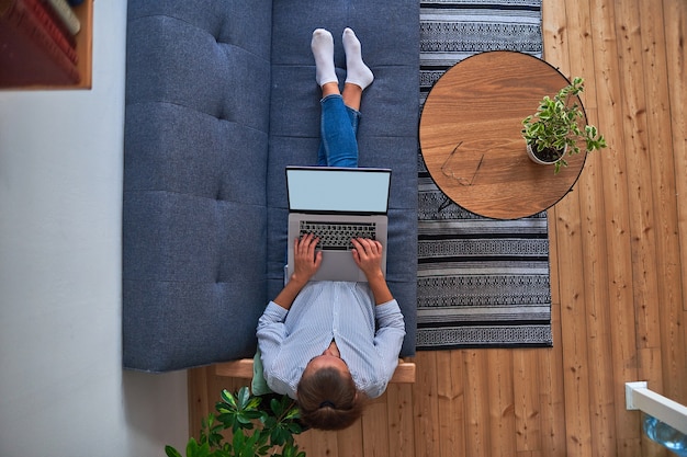 Mujer independiente sentada en el sofá y trabajando en línea en una computadora en un espacio de trabajo acogedor y acogedor con plantas de interior. Vista superior