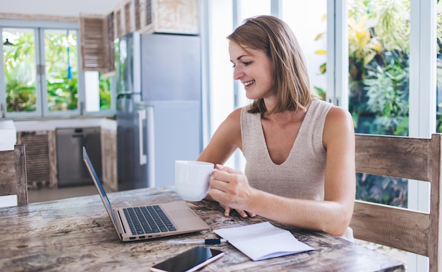 mujer independiente sonriente con una taza sentada en una mesa de madera en Bali Villa y trabajando en una laptop