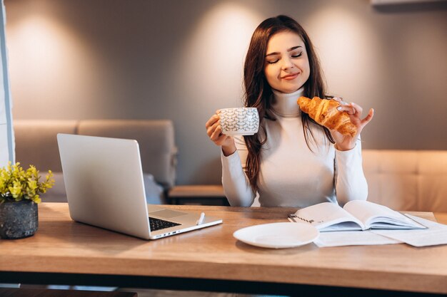 Mujer independiente usando laptop mientras está sentado en casa. Mujer joven sentada en la cocina y trabajando en equipo portátil. Bonita mujer bebiendo té café mientras trabaja.