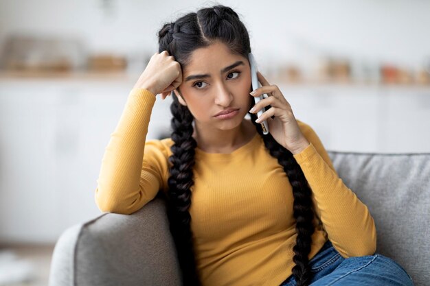 Mujer india aburrida hablando por celular mientras se sienta en el sofá en casa