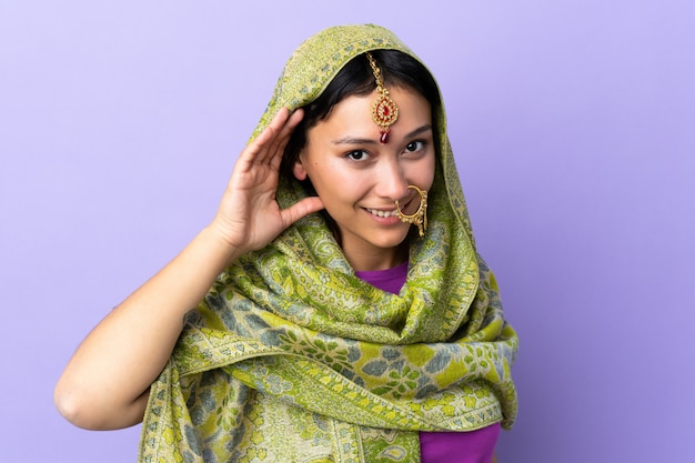 Mujer india aislada en púrpura escuchando algo poniendo la mano en la oreja