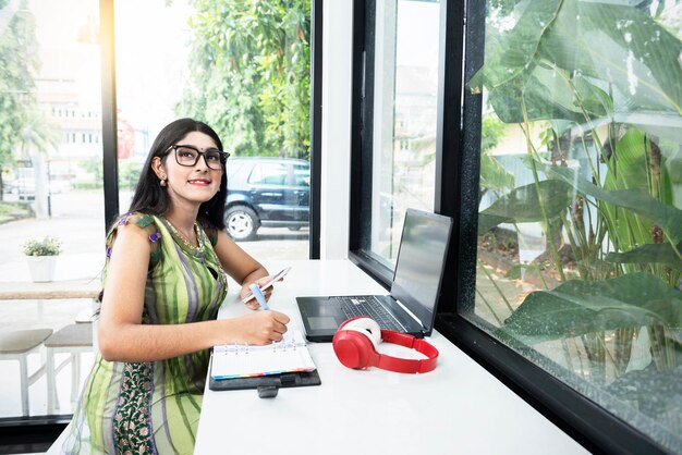 Mujer india con anteojos escribiendo algo en un cuaderno mientras sostiene un teléfono móvil con una computadora portátil en la mesa