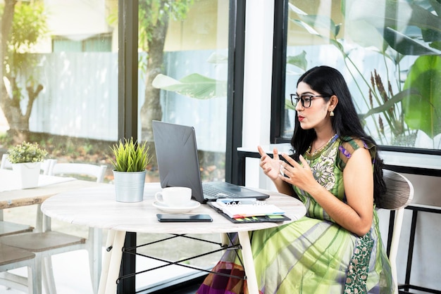 Mujer india con anteojos usando una computadora portátil con un cuaderno y un teléfono móvil con una taza de café en la mesa