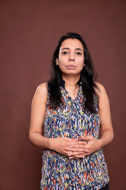 Mujer india con expresión facial neutra, retrato de dedos entrelazados. Señora tranquila, joven de pie con cara seria vista frontal estudio tiro medio sobre fondo marrón