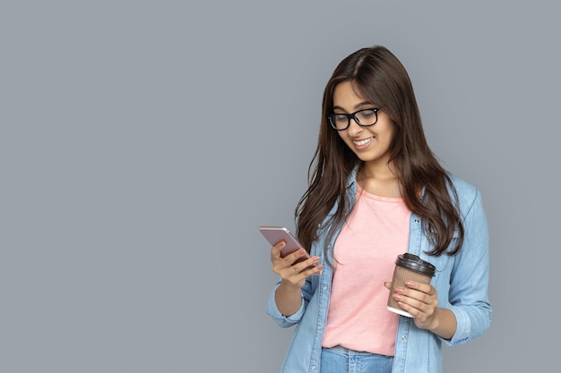 Foto mujer india joven adulta usando un teléfono inteligente sosteniendo una taza de café