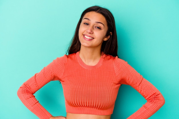 Mujer india joven aislada en azul confiado manteniendo las manos en las caderas.