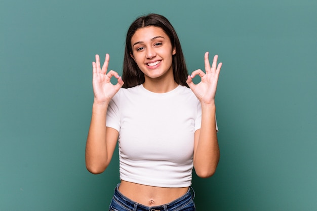 Mujer india joven aislada en la pared azul alegre y confiada que muestra el gesto aceptable.