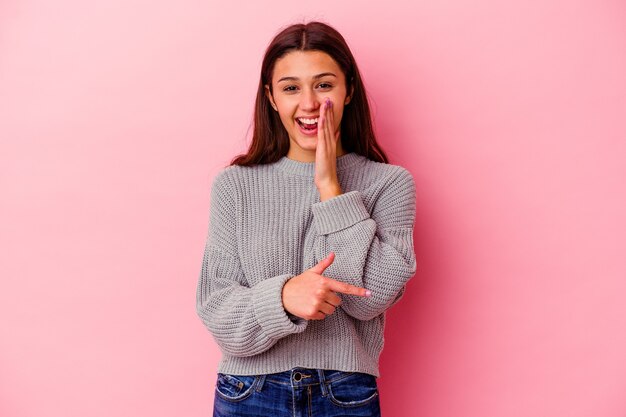 Mujer india joven aislada en la pared rosada diciendo un chisme, apuntando al lado que informa algo.