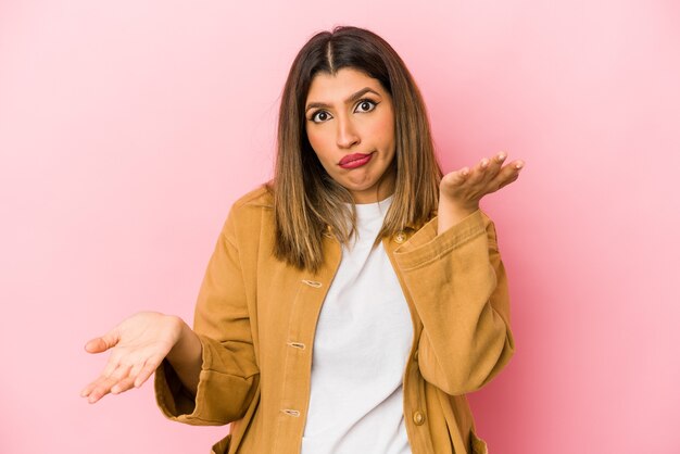 La mujer india joven aislada en la pared rosada hace escala con los brazos, se siente feliz y confiada.