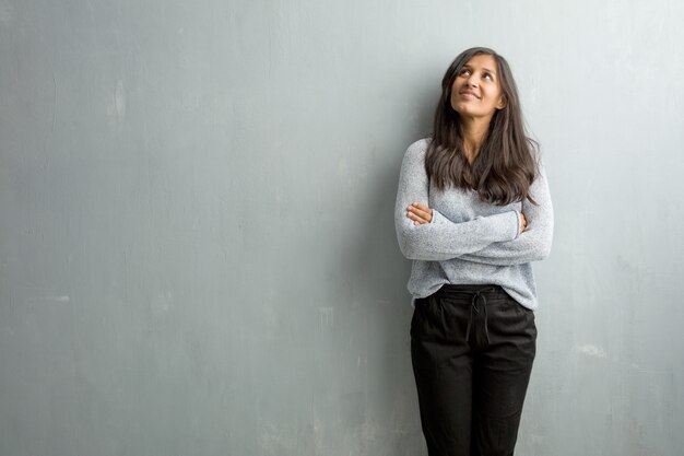 Mujer india joven contra una pared de grunge mirando hacia arriba, pensando en algo divertido y teniendo una idea, concepto de imaginación, feliz y emocionada