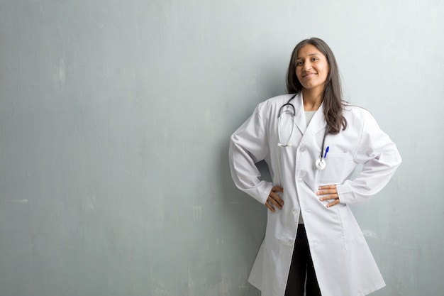 Mujer india joven del doctor contra una pared con las manos en las caderas, colocándose