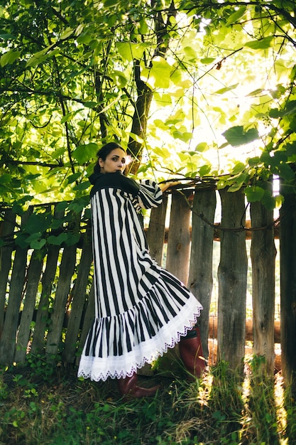 Mujer india joven y elegante vestida de blanco y negro sobre un fondo de árboles verdes y una valla de madera envejecida Mujer boho relajándose en el campo estilo de vida simple y lento Imagen atmosférica