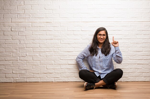 La mujer india joven se sienta contra una pared de ladrillo que muestra el número uno, símbolo de contar, concepto de matemáticas, confiado y alegre