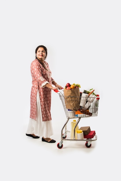 Mujer india de mediana edad con carrito de compras o carrito lleno de comestibles, verduras y frutas. Foto de cuerpo entero aislado sobre pared blanca