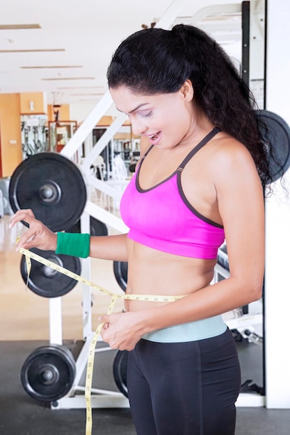 Foto la mujer india parece feliz mientras mide su cintura y está de pie en el gimnasio.