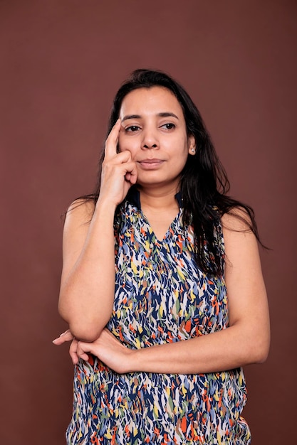 Mujer india pensativa mirando retrato de lado, modelo posando, sosteniendo el dedo en la cara. Persona pensativa de pie con expresión facial dudosa, plano medio del estudio de vista frontal