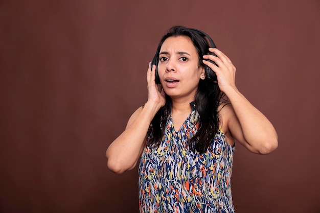 Mujer india quitándose los auriculares inalámbricos, escuchando música a todo volumen, mirando la cámara con una expresión facial confusa. Retrato de dama con auriculares, plano medio de estudio de vista frontal