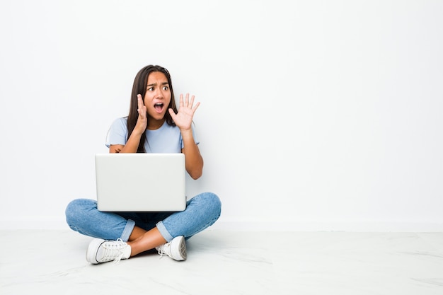 La mujer india de raza mixta joven que se sienta trabajando en la computadora portátil grita fuerte, mantiene los ojos abiertos y las manos tensas.